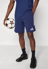 Σκούρο μπλε αθλητικό σορτς με λευκές ρίγες Adidas, κατασκευασμένο από απαλό ύφασμα. Το μοντέλο κρατάει μια ασπρόμαυρη ποδοσφαιρική μπάλα, φοράει μαύρα ποδοσφαιρικά παπούτσια.