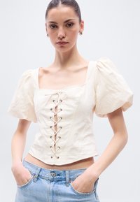 Ανοιχτό μπεζ κοντό τοπ με φουσκωτά μανίκια, τετράγωνο λαιμόκοψη και δέσιμο μπροστά με μεταλλικά στεφάνια. Συνδυάζεται με ανοιχτό μπλε τζιν παντελόνι.