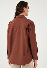 Brown button-up shirt with a collar, long sleeves, and side slits. Features a smooth texture and a slight drape at the back.