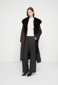 Καφέ μακρύ παλτό από σουέτ με μαύρο γιακά από γούνα, σε συνδυασμό με λευκή μπλούζα και γκρι παντελόνι με φαρδιά μπατζάκια, ολοκληρωμένο με κομψά μαύρα τακούνια.