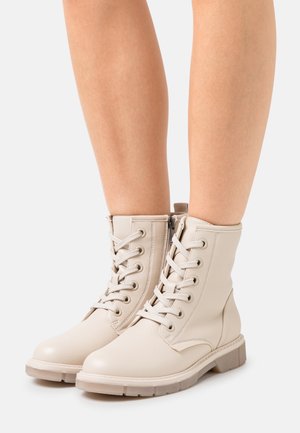 Hellbeige Schnürstiefeletten mit seitlichem Reißverschluss, getragen an nackten Beinen vor weißem Hintergrund.