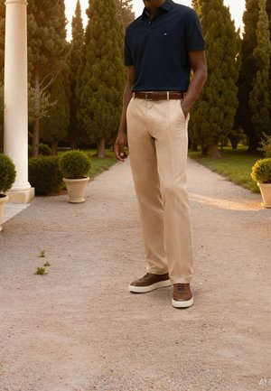 Uomo in piedi su un sentiero ghiaioso nel parco, che indossa una polo blu navy, pantaloni beige, cintura marrone e sneakers marroni, circondato da alberi alti e piante in vaso.