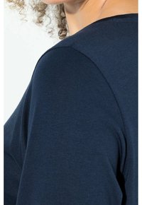 Eine Person mit lockigem Haar trägt ein eng anliegendes, dunkelblaues Oberteil, das die Schulter- und obere Brustpartie zeigt.
