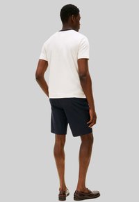 T-shirt da uomo a maniche corte in cotone color crema con dettagli blu navy; abbinata a pantaloncini blu navy scuro; modello di spalle, indossa mocassini marroni.