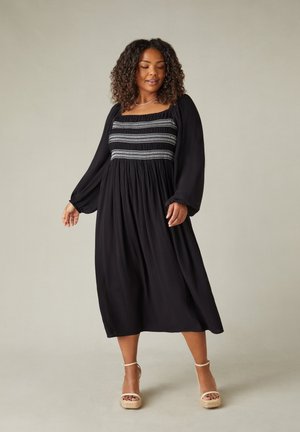 Schwarzes Maxikleid mit langen Ärmeln, ausgestattet mit einem gerafften Oberteil und textiler Oberfläche. Auffällige Raffung an der Taille, glatter Rock und ohne Muster.