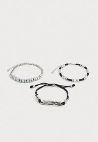Tres pulseras: una de cadena de metal plateado, una con cuentas plateadas sobre un cordón negro, y una de cordón negro con un centro de plata texturizada.