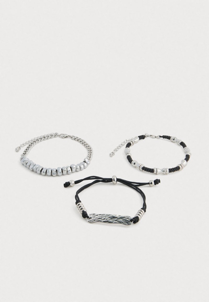 Tres pulseras: una de cadena de metal plateado, una con cuentas plateadas sobre un cordón negro, y una de cordón negro con un centro de plata texturizada.