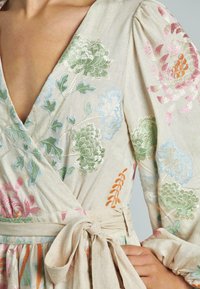 Besticktes Wickelkleid aus leichtem beigen Leinen mit floralen Mustern in Grün, Pink und Blau sowie einem gebundenen Gürtel in der Taille.