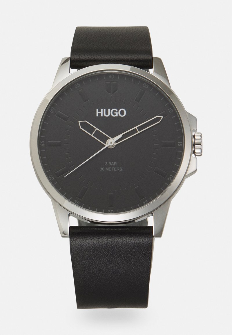 Montre-bracelet Hugo noire avec boîtier argenté, bracelet en cuir noir et cadran minimaliste affichant les aiguilles des heures, des minutes et des secondes.