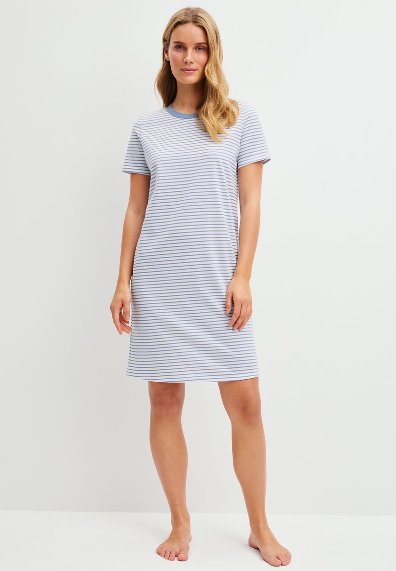 Jonge vrouw die blootsvoets staat en een witte jurk draagt tot op de knie met korte mouwen en dunne blauwe horizontale strepen.