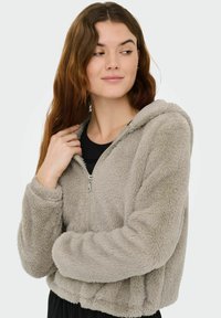 Sweat à capuche court en fausse fourrure grise avec fermeture à glissière à l'avant, capuche, poignets et ourlet côtelés, présentant une finition douce et texturée.