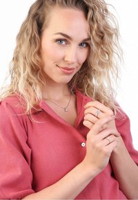 Cheveux blonds et bouclés ; portant une chemise rose à boutons. Collier en argent et deux bagues aux doigts. Gros plan, expression souriante, légèrement tourné à droite.