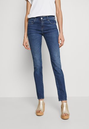 Frau trägt blaue Slim-Fit-Jeans, weißes T-Shirt und goldene Keilsandaletten mit offenen Zehen, stehend vor einem einfarbigen hellen Hintergrund.
