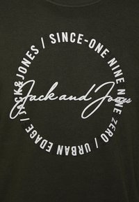 T-shirt di cotone verde scuro con una grafica circolare bianca che presenta la scritta "Jack and Jones" in corsivo e elementi testuali in font diversi.