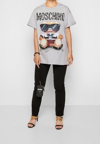 Grå oversized t-shirt med björngrafik som bär solglasögon, svarta byxor och svarta strappy klackar med guldkedjedetalj. Svart handväska.