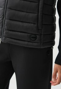 Schwarze gefütterte Weste mit steppdesign, Frontreißverschluss und runder Logopatch. Passt sich der Körperform an, kombiniert mit schwarzen, enganliegenden Leggings.