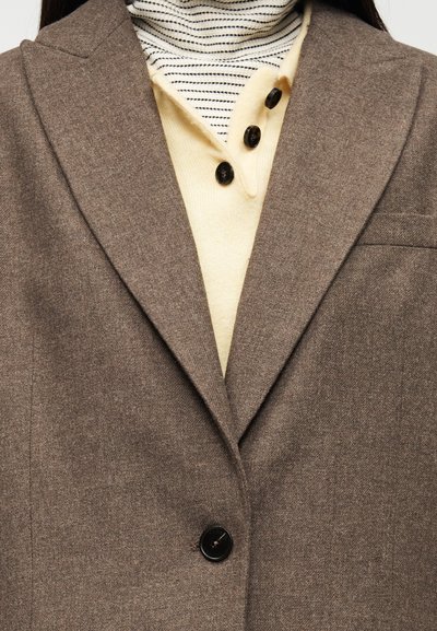 Καφέ μάλλινο σακάκι με ένα μαύρο κουμπί, με πέτο με εγκοπή και τσέπη στο στήθος, φορεμένο πάνω από ένα ανοιχτό κίτρινο τοπ με μαύρες ρίγες.