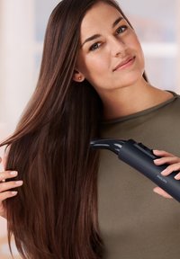 Outil de coiffure noir tenu par une personne avec de longs cheveux bruns lisses. L'outil possède une buse arrondie et des boutons de contrôle sur le côté.