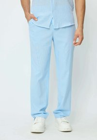 Homme portant un pantalon bleu clair, une chemise rayée bleu et blanche, des baskets blanches, se tenant avec une main dans la poche devant un fond uni.