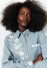 Chemise en denim bleu clair avec broderie florale blanche sur l'épaule gauche et la poche, dotée d'un col et d'une fermeture à boutons.