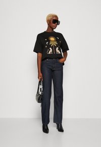 T-shirt grafica nera con design di sole e palma, jeans blu, stivaletti neri con tacchi, e una piccola borsa a tracolla con accenti circolari.