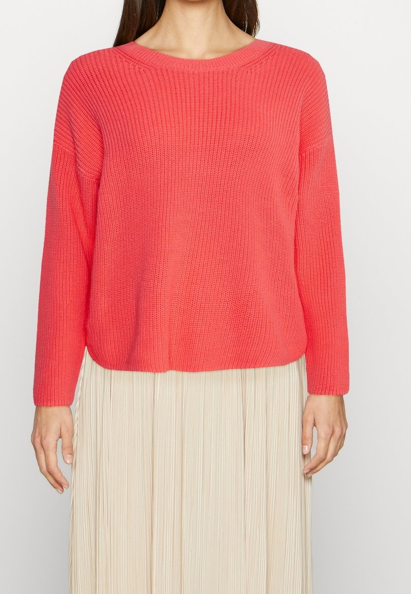Femme portant un pull en maille corail vif avec un col rond et des manches longues, associé à une jupe plissée beige.