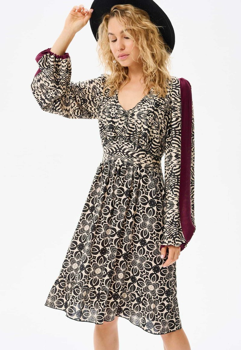 Bloemmotief jurk met een aansluitende lijfje en een uitlopende rok. Heeft lange mouwen met een diepe bordeauxrode accent en een V-hals.