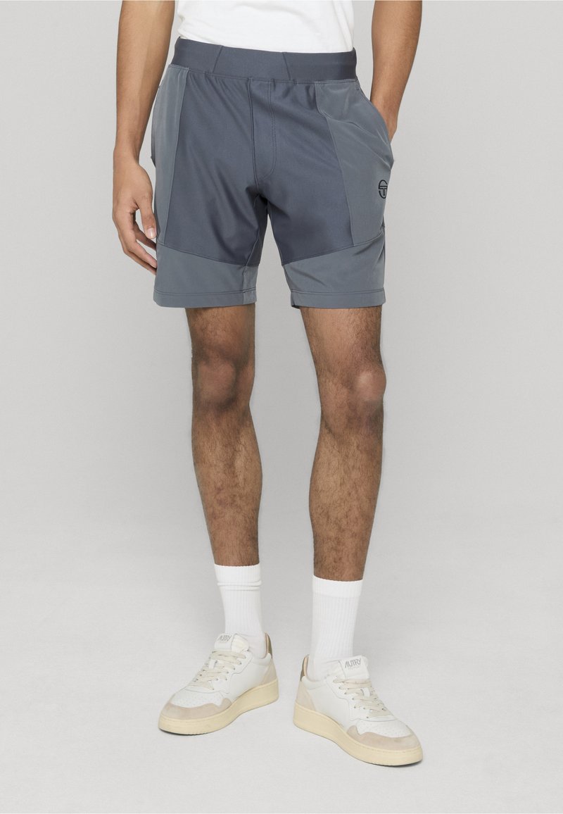 Homme portant un short de sport gris avec poches latérales, des chaussettes blanches montantes et des baskets blanches sur un fond clair uni.