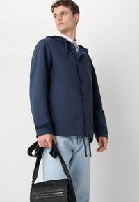 Navy lightweight jacket with a hood, button closure, and textured cuffs; paired with light blue jeans and a black crossbody bag.