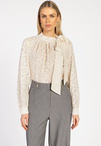 Vrouw in doorschijnende crèmekleurige bloemenkant blouse met grote strik om de hals en hooggetailleerde grijze broek tegen een witte achtergrond.