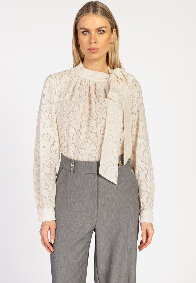 Vrouw in doorschijnende crèmekleurige bloemenkant blouse met grote strik om de hals en hooggetailleerde grijze broek tegen een witte achtergrond.