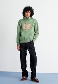 Hoodie verde com um logo impresso, combinado com jeans largos pretos e sapatos castanhos com atacadores sobre uma superfície de carpete azul.