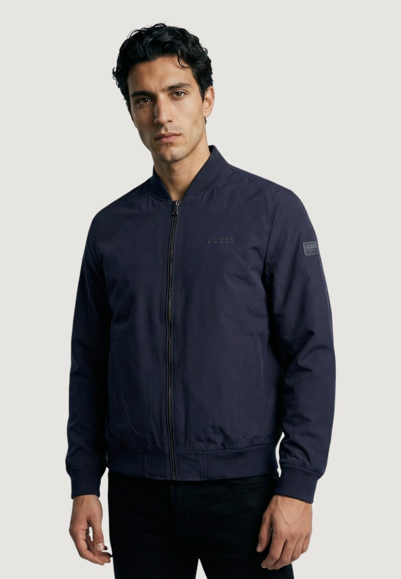 Blouson aviateur bleu marine avec fermeture éclair à l'avant, poignets côtelés, logo GUESS sur la poitrine et poches latérales ; fabriqué en tissu lisse et léger.