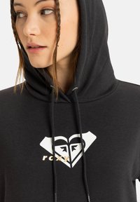 Felpa nera con cappuccio e coulisse, decorata da un logo a forma di cuore bianco e oro sul petto. Tessuto morbido con una vestibilità rilassata.