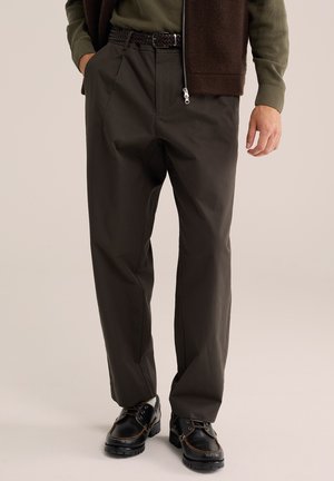 Homme portant un pantalon droit marron foncé avec une ceinture tressée, un pull vert, un gilet foncé et des chaussures noires à lacets.