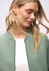 Giacca bomber verde trapuntata con motivo a spina di pesce, dotata di chiusura con zip e collo arrotondato, indossata sopra una maglia bianca.