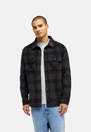 Jeune homme portant une veste à carreaux noire et gris foncé boutonnée sur un t-shirt gris clair et un jean bleu, debout devant un fond uni.