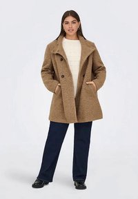 Moteris vilki smėlio spalvos tekstūruotą dvigubą paltą ant balto megztinio, tamsių kelnių ir juodų batų, stovinti su rankomis kišenėse.