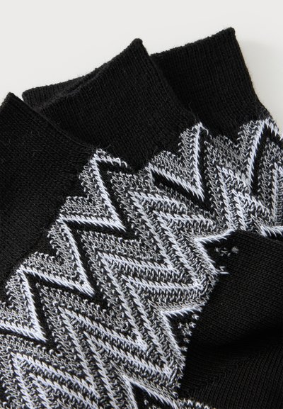 Chaussettes en maille noires avec un motif en zigzag blanc et gris et des bord-côtes noirs unis superposés sur un fond blanc.