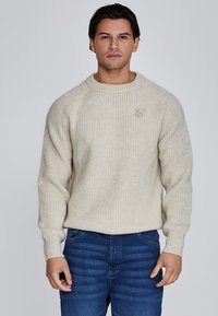 Beżowy sweter z dzianiny o ribbowanej fakturze, okrągłym dekolcie i małym haftowanym logo na piersi. Noszony z niebieskimi dżinsami.