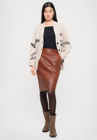 Cardigan lavorato a maglia beige con bottoni e ricami, top nero, gonna a tubino in pelle marrone, collant scuri e stivaletti marroni.