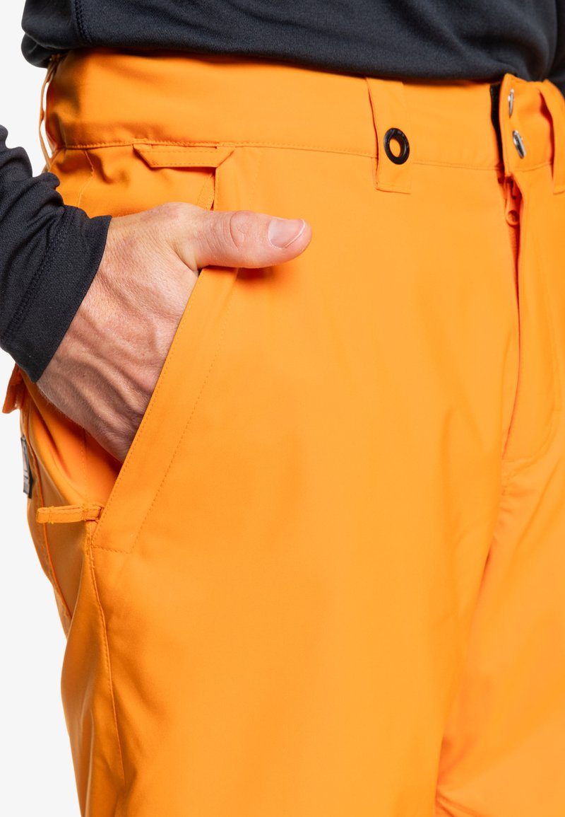 Pomarańczowe techniczne spodnie o gładkiej fakturze, z bocznymi kieszeniami i widocznym zapięciem na guzik. Ręka umieszczona w kieszeni.