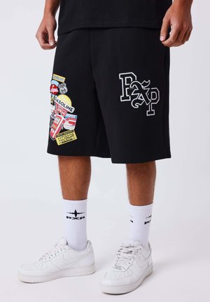 Homme portant un short noir orné de patches colorés de station-service, des chaussettes blanches avec des logos noirs et des baskets blanches, sur un fond uni.