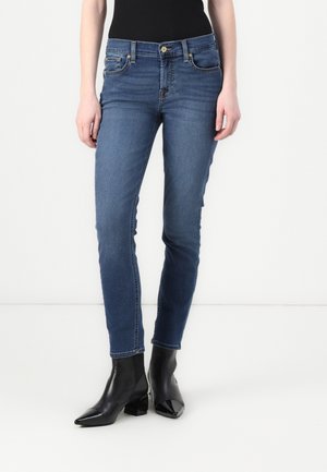Kvinde iført tætsiddende blå jeans og sorte ankelstøvler med hæl, stående mod en hvid baggrund.