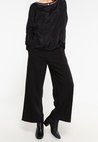 Blouse noire à manches longues avec un col rond orné de garniture embellie, associée à un pantalon noir à jambes larges et des bottines noires.