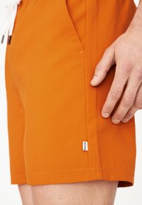 Oranžové plavky vyrobené z lehkého materiálu. Obsahují boční kapsy, stahovací pas a malou značkovou nášivku na lemu.