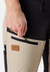 Beige och svarta friluftsbyxor har en ficka med dragkedja och en läderlogg. Tyget verkar hållbart och flexibelt, lämpligt för aktiviteter.