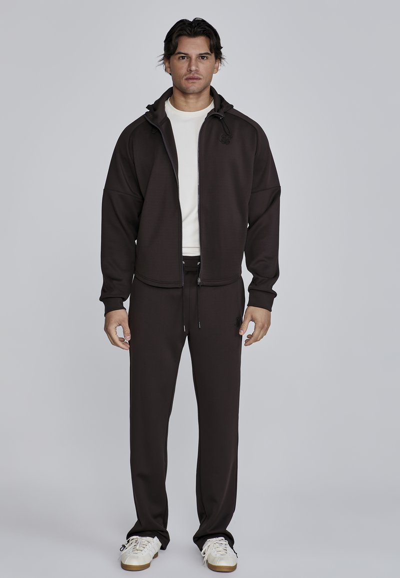 Conjunto deportivo de color marrón oscuro que incluye una chaqueta con cremallera y capucha, pantalones jogger a juego y zapatillas blancas con acetatos en beige.