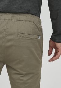 Licht olijfgroene broek met een elastische tailleband, met een achterzak en een klein merklabel op de zak. Glad materiaal, aansluitend ontwerp.