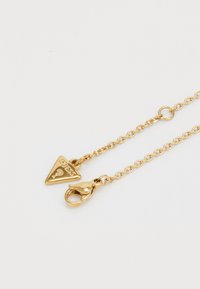 Collier en chaîne dorée avec un charm triangulaire gravé "GUESS", et un fermoir mousqueton. Texture lisse, design minimal.